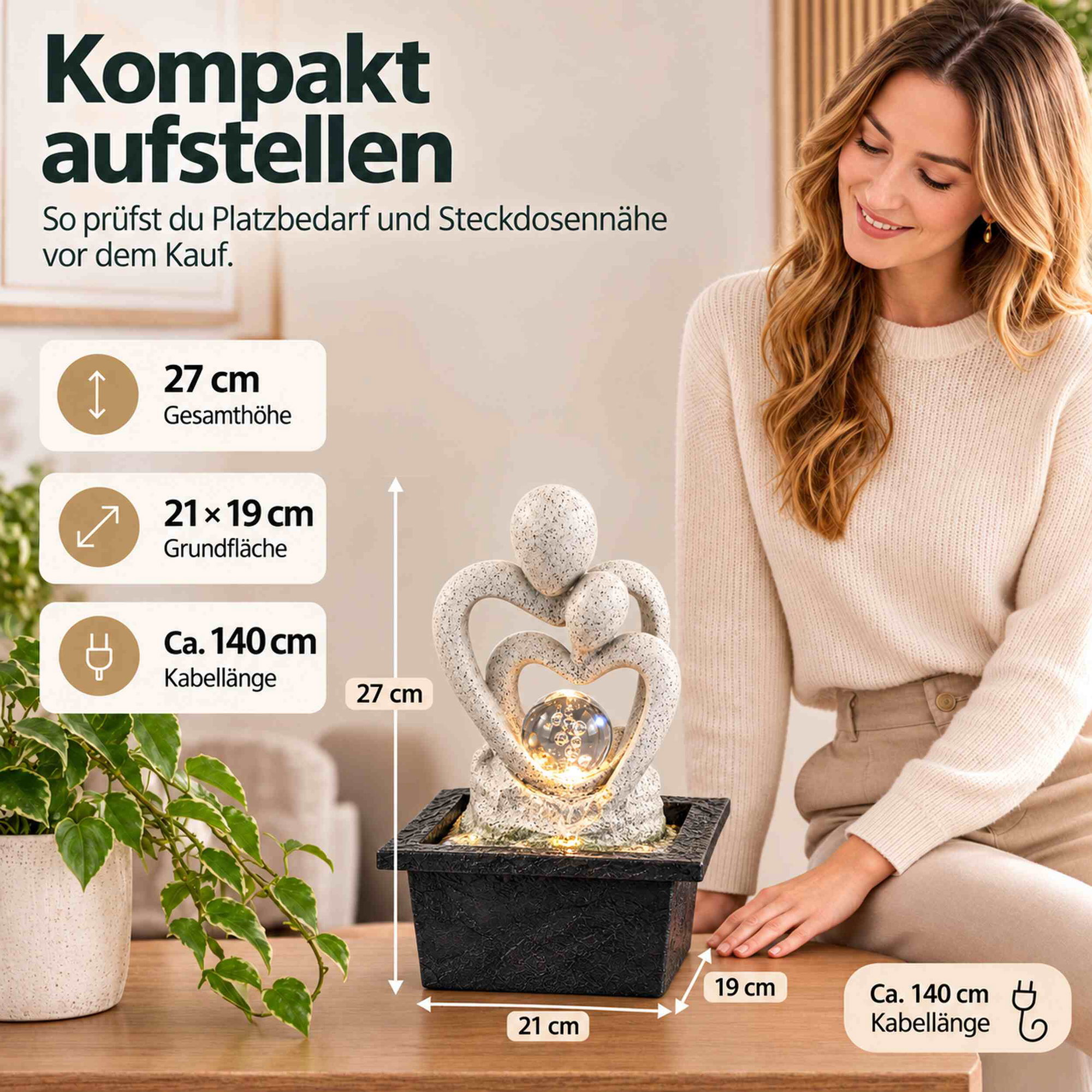 Zimmerbrunnen mit LED-beleuchteter Glaskugel und Paar-Skulptur in Granitoptik auf einem Tisch, daneben eine lächelnde Frau und eine Pflanze, mit Maßangaben 27 cm Höhe, 21 x 19 cm Grundfläche und 140 cm Kabellänge